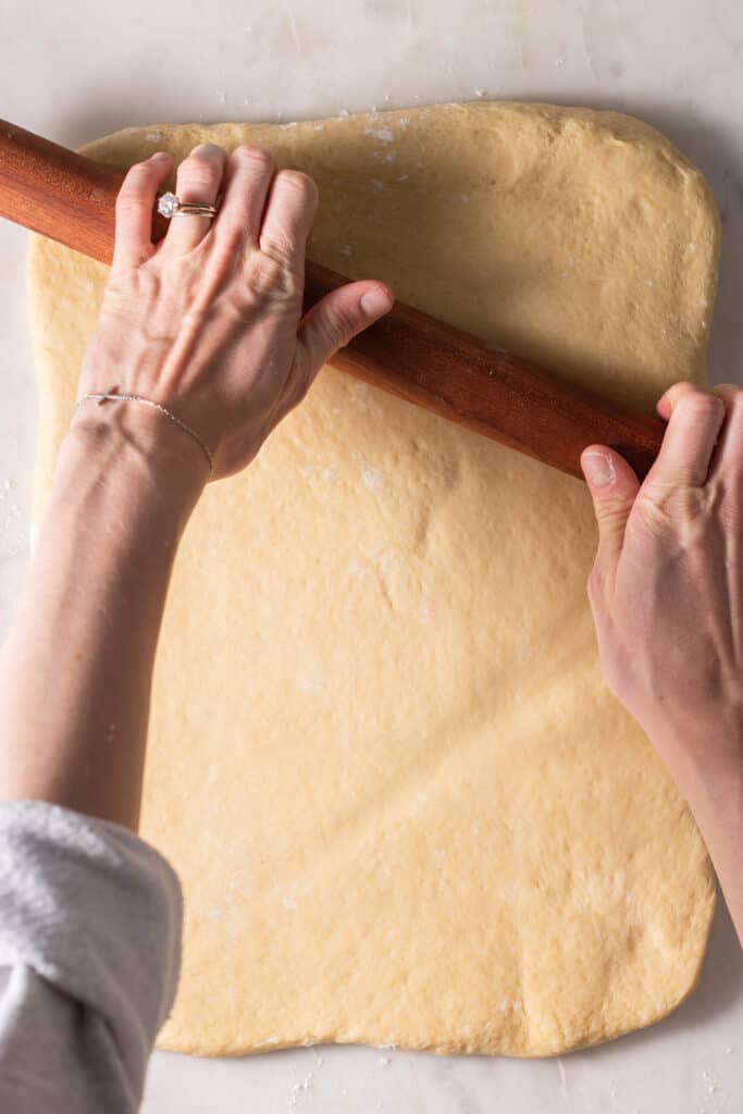 hands rolling out cinnamon roll dough on a floured surface with a rolling pin