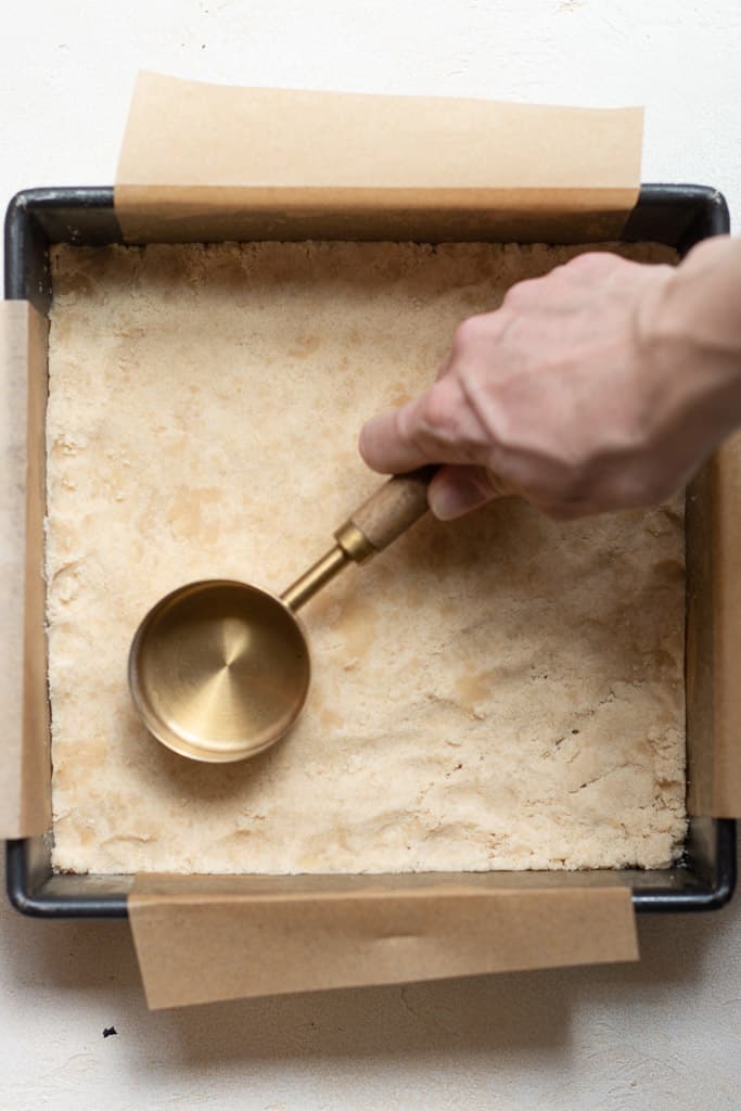 hand patting shortbread crust into the bottom of a pan using the bottom of a measuring cup.