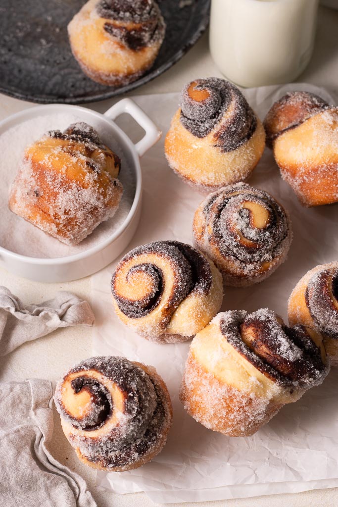 multiple chocolate buns on a board with one being rolled in sugar.