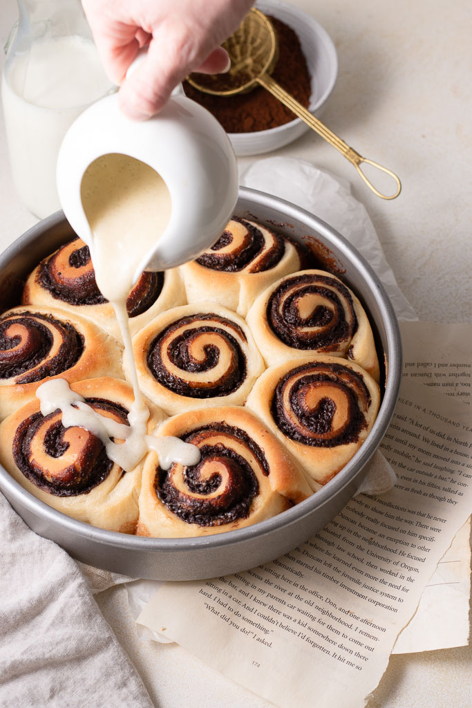 side angle shot of a hand pouring a glaze overtop of chocolate cinnamon rolls.